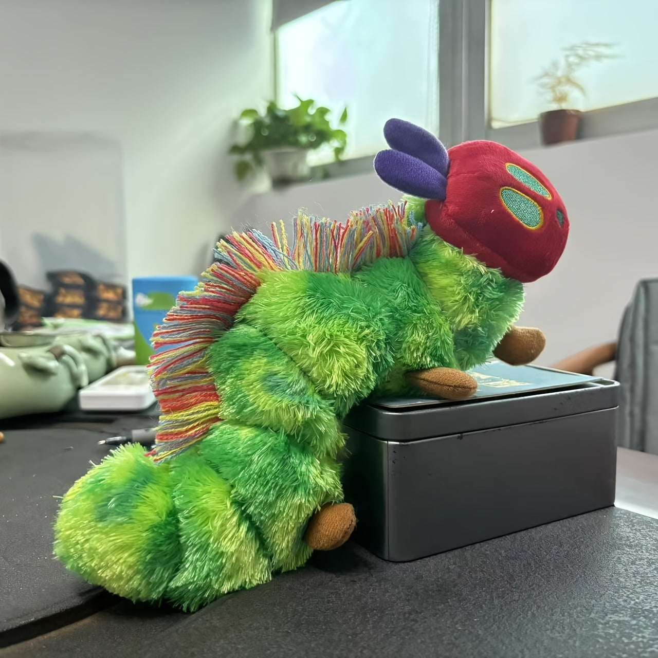 Green caterpillar plush toy with colorful yarn spikes and a red head sitting on a black box